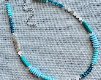 Op de afbeelding: Een turquoise en zilveren kralenketting met een verloop van kleuren. De ketting is gemaakt met ronde kralen in verschillende tinten blauw en turquoise, afgewisseld met ronde zilveren kralen. De ketting heeft een zilveren sluiting.
