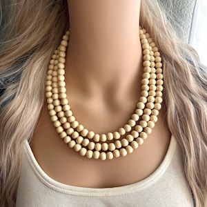 May include: A triple-strand necklace composed of smooth, round, light beige wooden beads. The beads are uniform in size and color, strung together to form three distinct, layered circular rows that rest against the neck area.