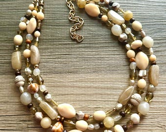 May include: A multi-strand necklace featuring a variety of beads in cream, beige, and brown tones. The necklace includes round, oval, and irregular-shaped beads, with gold-colored metal accents and a clasp.