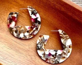May include: A pair of large, brown, white, and pink hoop earrings with a unique, irregular, marbled pattern. The earrings are made of a lightweight, durable material.