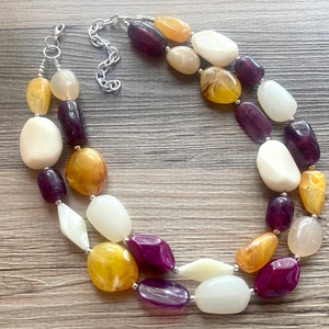 Peut inclure: Un collier à double rang de perles ovales, épaisses et de grande taille, dans des tons de blanc, violet et jaune. Les perles sont en matériau lisse et poli.