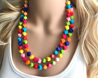 Puede incluir: Un collar multicolor hecho de cuentas cuadradas. Las cuentas están dispuestas en un patrón de arcoíris, con cada cuenta de un color diferente. El collar está en capas, con dos hileras de cuentas.