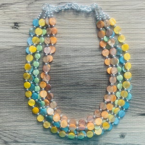 May include: A multi-strand necklace featuring large, iridescent, round beads in shades of yellow, blue, orange, and green. The beads are strung together with silver-colored metal accents and a silver chain. The necklace is a statement piece.