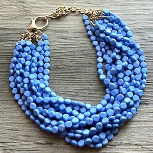 May include: A multi-strand necklace featuring light blue, round beads. The necklace has a gold-colored chain and clasp. The beads are arranged in multiple rows, creating a layered effect. This statement necklace is a fashion accessory.