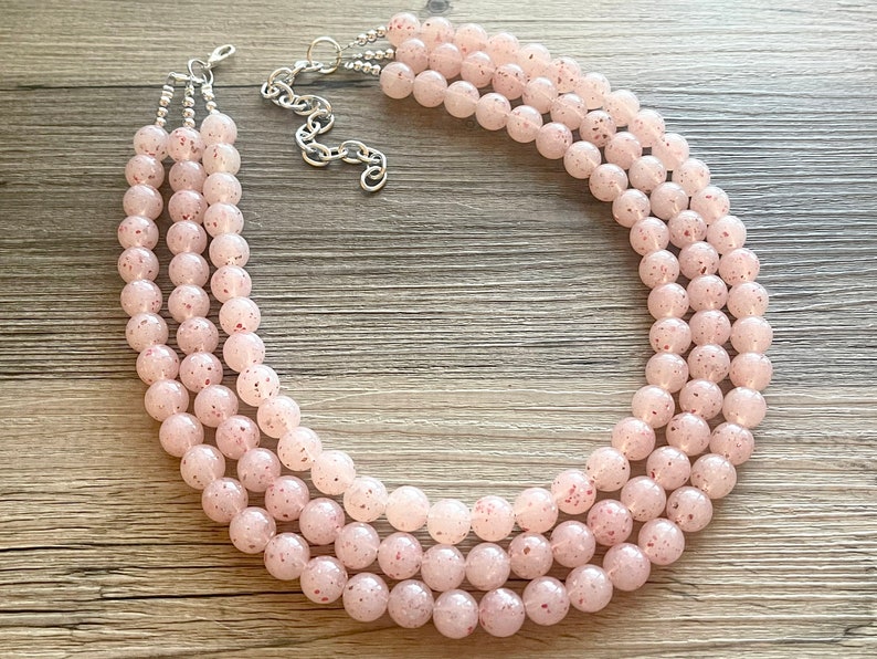 May include: A three-strand necklace made of light pink beads with a speckled texture. The necklace has a silver clasp.
