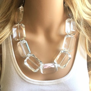 May include: A clear, chunky statement necklace with large, rectangular, faceted beads. The necklace is strung on a clear cord and is displayed on a person wearing a white tank top. The necklace has a modern, bold design.