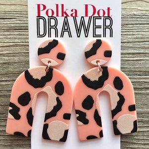 Könnte beinhalten: Rosa und schwarze Statement-Ohrringe mit Leopardenmuster in abgerundeter Bogenform. Die Ohrringe sind aus Polymer Clay gefertigt und haben einen silbernen Haken.