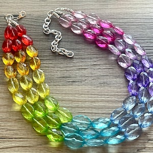 Op de afbeelding: Een meerstrengs ketting met een regenboogverloop van facetgeslepen ovale kralen. De kleuren gaan van rood naar geel, groen, blauw en paars. De ketting heeft een zilverkleurige ketting en sluiting.