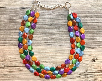 Puede incluir: Un collar de múltiples hilos hecho de cuentas ovaladas de colores. Las cuentas están dispuestas en un patrón de arcoíris, con tonos de rojo, naranja, amarillo, verde, azul y morado.