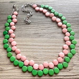 Könnte beinhalten: Eine zweireihige Halskette mit abwechselnd rosa und grünen runden Perlen. Die Halskette ist aus Kunststoff und hat einen silbernen Verschluss.