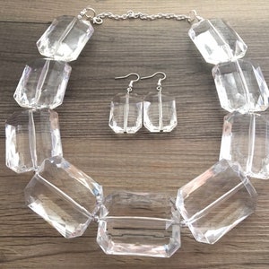 May include: A clear, faceted crystal necklace and matching earrings set. The necklace features large, rectangular crystal beads strung on a silver chain. The earrings are smaller, rectangular crystals with silver hooks. The jewelry set is displayed on a wooden surface.