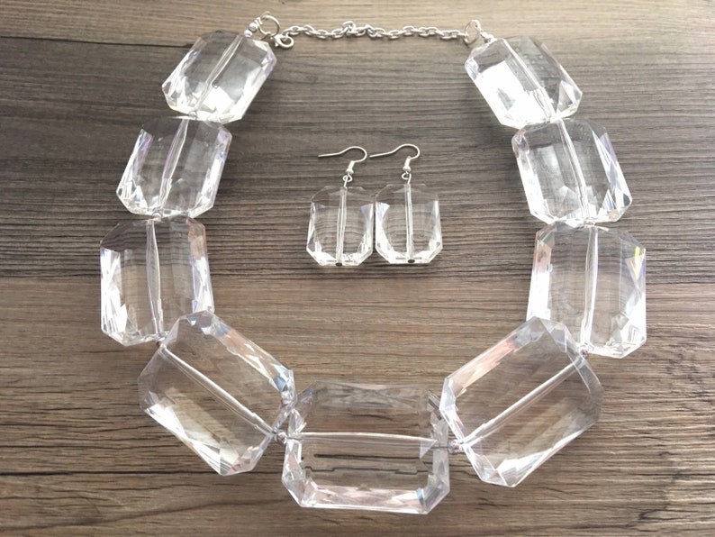 May include: A clear, faceted crystal necklace and matching earrings set. The necklace features large, rectangular crystal beads strung on a silver chain. The earrings are smaller, rectangular crystals with silver hooks. The jewelry set is displayed on a wooden surface.