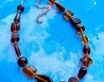 May include: A brown glass beaded necklace with gold accents. The beads are various shapes and sizes, including round, oval, and square.