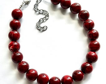 Può includere: Una collana composta da una serie di grandi perle rotonde rosso lucido con sottili motivi marmorizzati. Le perle sono infilate e collegate a una catena in metallo color argento con chiusura a moschettone e una catenella di estensione regolabile.
