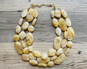 May include: A multi-strand necklace made of large, irregular, light brown and white stones. The necklace is made of three strands of stones, and the stones are connected by gold-colored metal links.