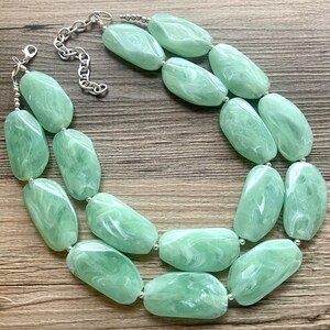 May include: A double-strand necklace made of large, light green, oval-shaped beads with a swirling pattern. The beads are connected by silver-toned metal.