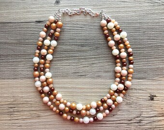 May include: A multi-strand necklace made of brown, beige, and white beads. The necklace is made of three strands of beads, each with a different color. The beads are all round and have a smooth finish.