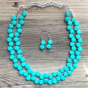 May include: A turquoise blue necklace and matching earrings set. The necklace is made of round beads and has a silver chain closure. The earrings are dangle style with two round beads.