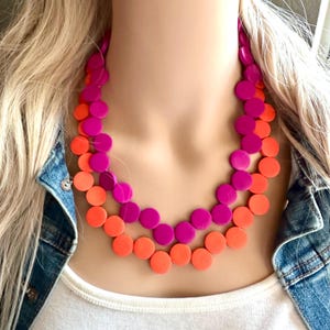 May include: A two-strand necklace featuring large, round beads in vibrant colors. The top strand is a deep magenta, while the bottom strand is a bright orange. The necklace is displayed on a mannequin.