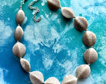 May include: A necklace featuring large, oval-shaped beads in shades of beige and brown, strung together with a silver-colored chain. The beads have a cracked, textured appearance. The necklace is displayed against a blue and teal background.