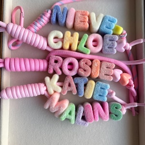 May include: Five personalised keychains featuring fuzzy, multi-coloured letter beads spelling out the names NIEVE, CHLOE, ROSIE, KATIE, and MALINS. Each keychain is attached to a pink cord with a matching pink clasp and small heart-shaped beads.