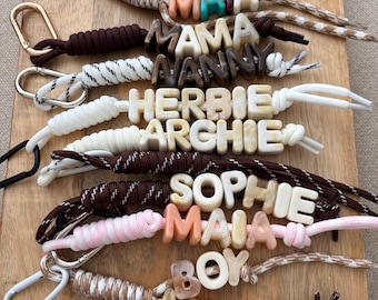 May include: A collection of personalised keychains featuring braided cords in various colours and patterns, each attached to a metal carabiner. The keychains are customised with individual letter beads in marbled, solid, and translucent finishes.