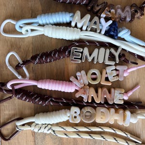 May include: A collection of personalised keychains featuring braided nylon cords in various colours including brown, white, light blue, and pink. Each keychain includes a metal or acrylic clasp and letter beads spelling names like MAMA, MAYA, EMILY, VIOLET, ANNE, and BODHI.