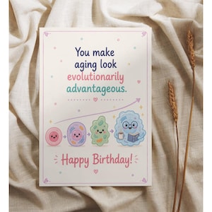 May include: A rectangular greeting card with a cream background featuring the text "You make aging look evolutionarily advantageous." and "Happy Birthday!". Below the text, four cartoon cells show an evolutionary progression, ending with a blue cell holding a book and a mug.