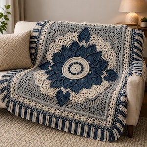 Op de afbeelding: Een rechthoekige gehaakte deken met een groot centraal mandala-ontwerp in diep marineblauw en crème. Het ingewikkelde patroon bevat gelaagde bloemblaadjes, cirkelvormige texturen en een decoratieve rand afgewerkt met lange, afwisselende franjes.