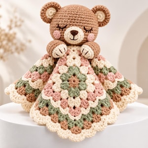 May include: A crocheted teddy bear security blanket featuring a brown bear head with closed eyes and pink cheeks. The attached blanket is made of a multi-coloured granny square pattern in shades of cream, sage green, and dusty rose with a scalloped edge.