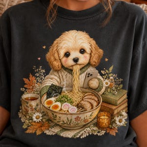 May include: A charcoal gray t-shirt featuring a graphic of a fluffy light brown puppy wearing a beige sweater, eating a bowl of ramen with chopsticks while holding a smartphone. The scene includes a mug, stacked books, and flowers.