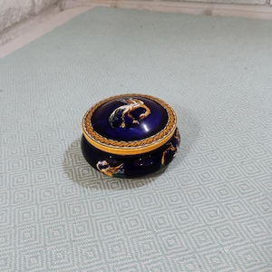 May include: A round, dark blue ceramic trinket box with a lid. The lid features a central painted bird illustration surrounded by a yellow and orange rope-style border. The base of the box also displays painted bird motifs in orange and white.