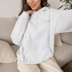 May include: A white hooded sweatshirt featuring a front kangaroo pocket and long sleeves. The chest area displays the text L&D embroidered in a light red outline font. The garment includes a matching white drawstring at the neckline.