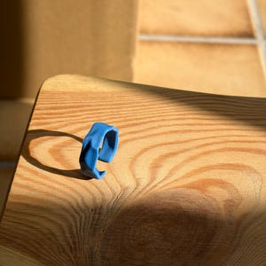 Op de afbeelding: Een felblauwe ring met een open bandontwerp en een getextureerd, ongelijkmatig oppervlak. De ring heeft een matte afwerking en een licht onregelmatige, organische vorm die een opvallend silhouet creëert en de gedurfde kleur benadrukt.