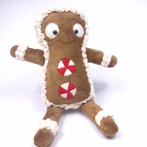 May include: A brown gingerbread man plush toy with white frosting trim and red and white candy decorations. The toy has a smiling face with black eyes and a brown nose.