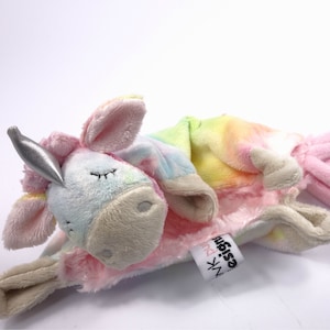 May include: A plush rainbow unicorn toy with a silver horn. The toy is lying on its side with its head resting on a pink blanket. The blanket has the text "design" and "pink" on it.