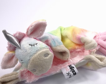 Puede incluir: Un juguete de peluche de unicornio arcoíris con un cuerno plateado. El juguete está acostado de lado con la cabeza apoyada en una manta rosa. La manta tiene el texto "design" y "pink" en ella.