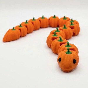 May include: A segmented toy shaped like a snake, constructed from a series of connected orange pumpkin figures with green stems. The head segment features two black eyes and small white fangs, creating a long, flexible, and articulated creature.