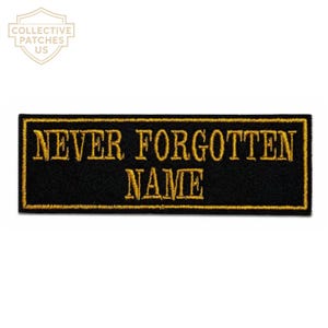 May include: A rectangular black embroidered patch with a gold border. The text "NEVER FORGOTTEN" is stitched in gold thread on the top line, and the word "NAME" is stitched in gold thread on the bottom line, centred below the first line.