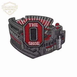 Peut inclure: Un écusson brodé représentant un stade dans des tons de gris, rouge et blanc. Le motif présente un octogone rouge proéminent avec le texte THE O SHOE en blanc, et une tour verticale sur le côté droit avec le nombre 1922 brodé en blanc.