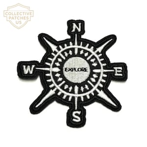 Puede incluir: Un parche bordado en blanco y negro con un diseño de rosa de los vientos. El círculo central contiene la palabra EXPLORE en letras negras. Los puntos cardinales N, S, E y W están situados en negro en las puntas de la brújula.