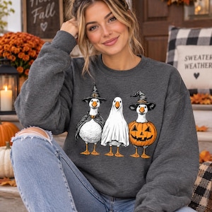 May include: A dark gray crewneck sweatshirt featuring a graphic of three white geese dressed for Halloween. One goose wears a black witch hat and cape, the middle goose is draped in a white ghost sheet, and the third goose wears a pumpkin costume.