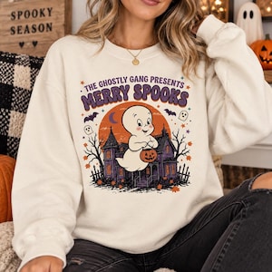May include: A cream-colored crewneck sweatshirt featuring a graphic print with the text THE GHOSTLY GANG PRESENTS MERRY SPOOKS. The design includes a white ghost holding a pumpkin bucket in front of a purple haunted house against an orange moon.