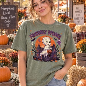 May include: A sage green short-sleeve t-shirt featuring a graphic print of a ghost holding a jack-o'-lantern in front of a haunted house. The text "FRIENDLY SPOOKS" is printed in purple above the illustration, which includes bats and a moon.