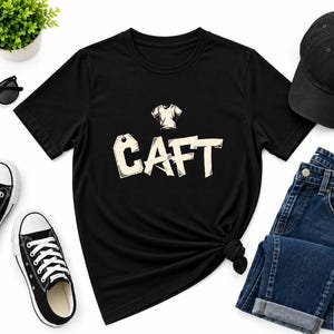 May include: A black short-sleeve t-shirt featuring a graphic of a small t-shirt icon above the text CAFT in a stylized, blocky font. The shirt is displayed alongside black sunglasses, a black baseball cap, and dark blue denim jeans.
