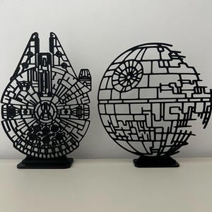 May include: Two black, flat, laser-cut decorative figures standing on rectangular bases. One figure is shaped like the Millennium Falcon spaceship, and the other is shaped like the Death Star space station, both featuring intricate geometric cutouts.