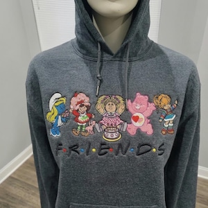 May include: A dark gray hooded sweatshirt featuring an embroidered graphic of five cartoon characters including a Smurf, Strawberry Shortcake, a Cabbage Patch Kid, a Care Bear, and Rainbow Brite, with the word FRIENDS embroidered below.
