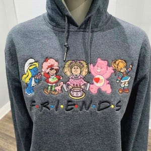 Puede incluir: Una sudadera con capucha gris carbón que presenta personajes de dibujos animados bordados, incluyendo un Pitufo, Rosita Fresita, un Cabbage Patch Kid, un Osito Cariñosito y Rainbow Brite. La palabra FRIENDS está bordada debajo en negro.