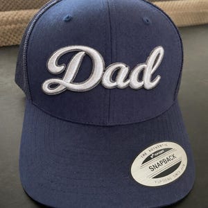 May include: A navy blue baseball cap with a mesh back and a curved brim. The front features the word "Dad" embroidered in white script with a grey outline. A circular sticker on the brim reads "THE AUTHENTIC YUPOONG SNAPBACK SINCE 1974".
