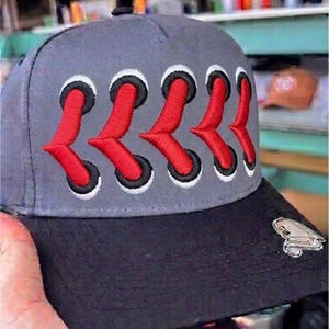 May include: A gray baseball cap featuring a black brim and a prominent embroidered design on the front panel. The design consists of five pairs of black-rimmed eyelets connected by red, V-shaped stitching. A small silver-toned metal frog emblem is attached to the right side of the black brim.